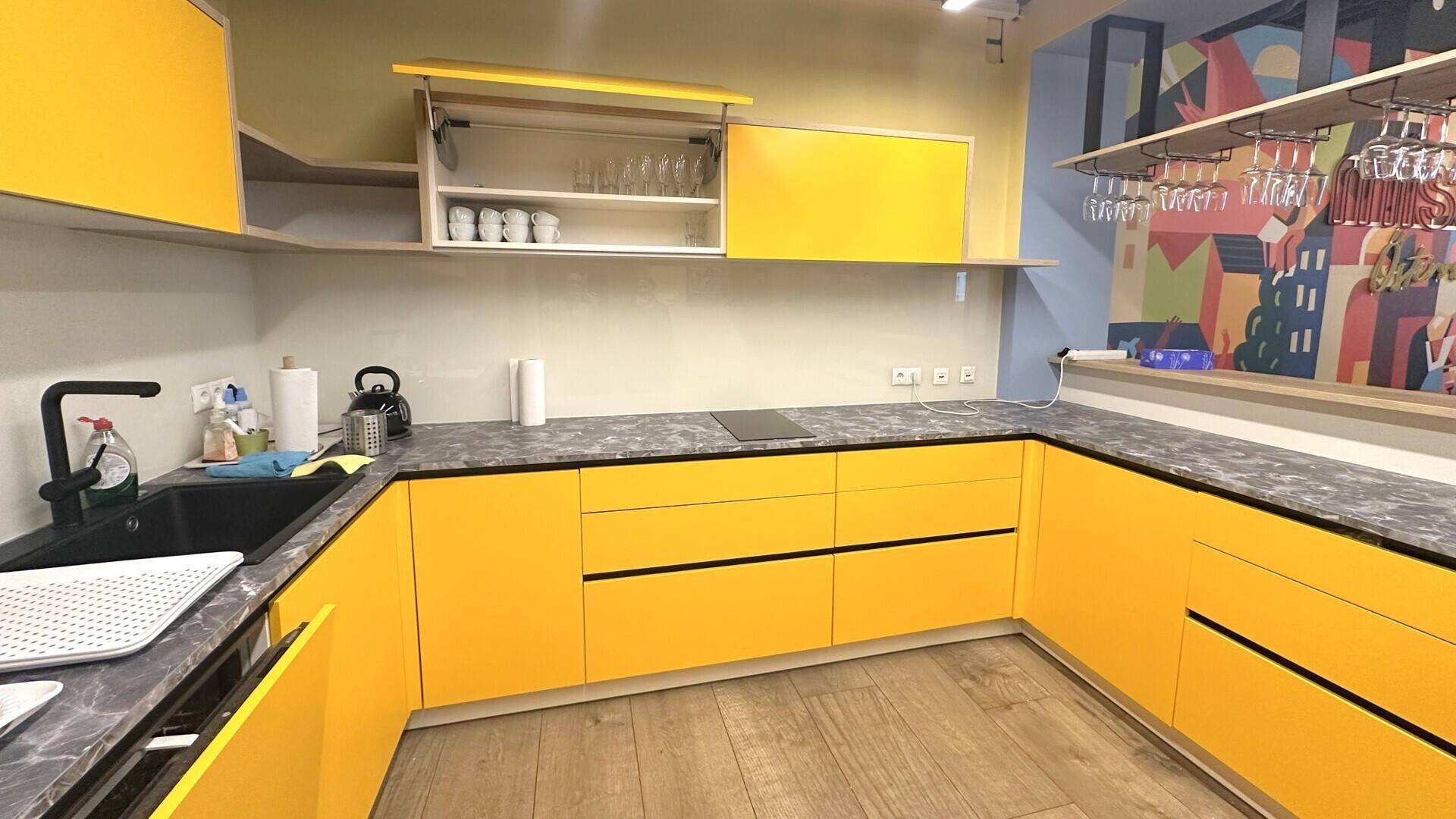 Moderne gelbe Küche mit dunklen Arbeitsplatten, Induktionskochfeld und bunten Wanddetails im Hintergrund.