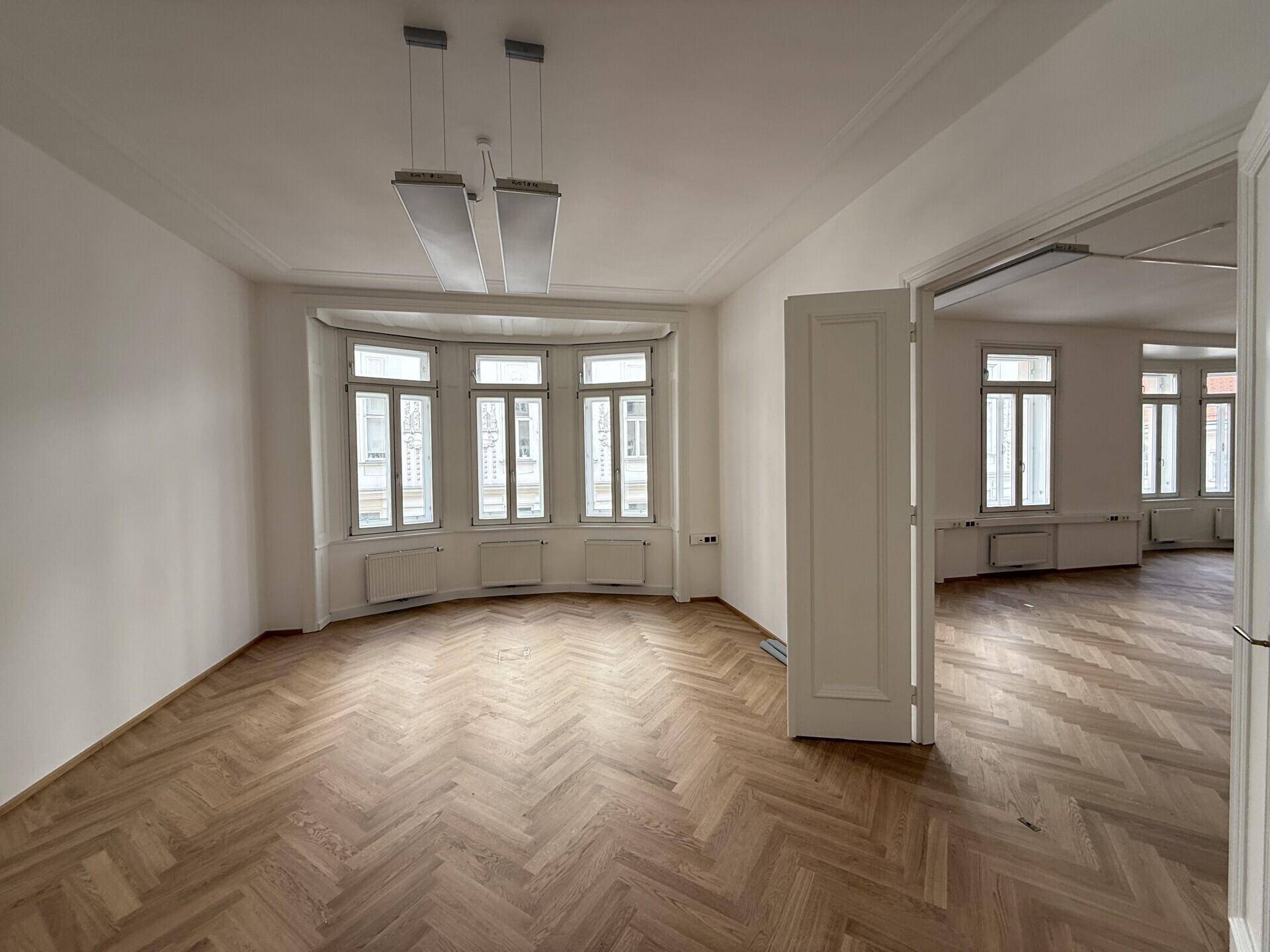 Büro mit Erkerfenstern, Fischgrätparkett und Deckenleuchten, das viel Tageslicht und Stadtblick bietet.