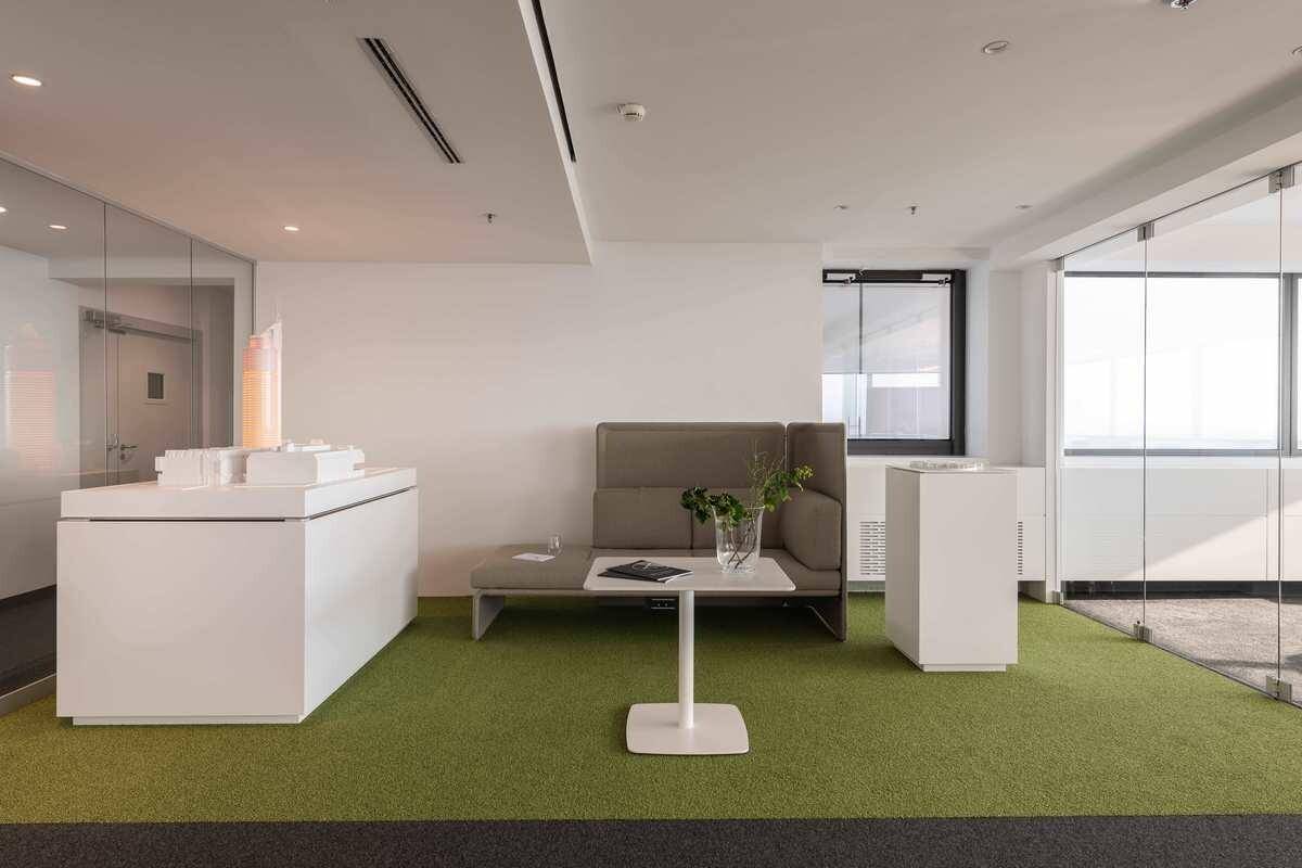 Heller Bürobereich mit einem Sofa, einem Couchtisch und einem grünen Teppichbereich, umgeben von Glaswänden.
