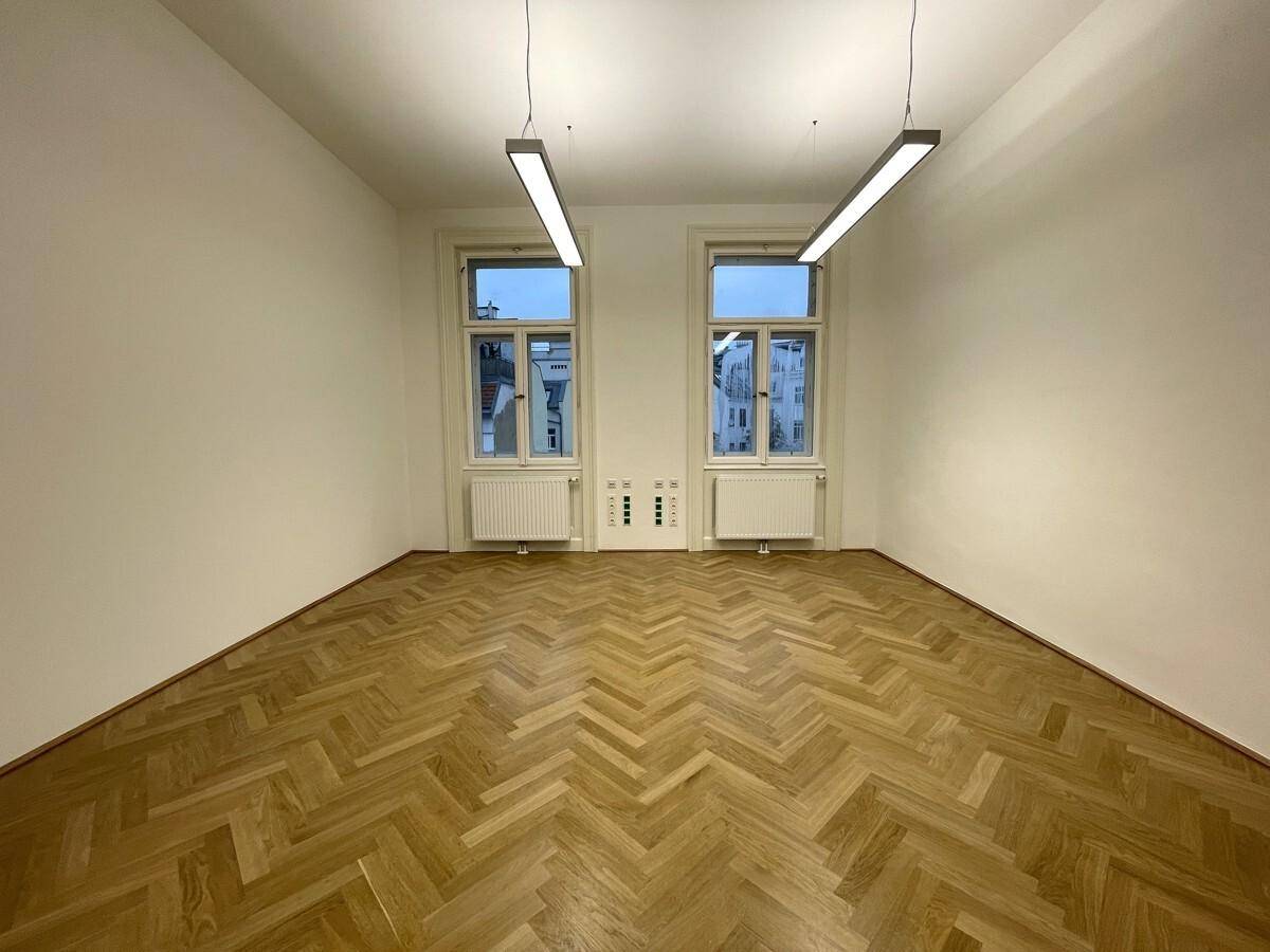 Geräumiger Büroraum mit Fischgrätparkett und zwei hohen Fenstern, die für eine helle Arbeitsumgebung sorgen.