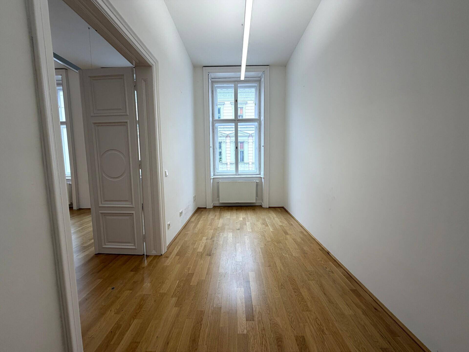 Schmaler Raum mit Holzboden, einem großen Fenster und weiß gestrichenen Wänden.