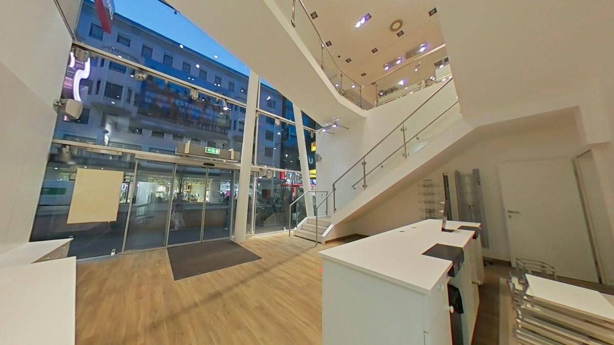 Geräumiger Verkaufsraum mit großen Glasfronten, Holzboden und einer modernen Treppe, die zum oberen Bereich führt.