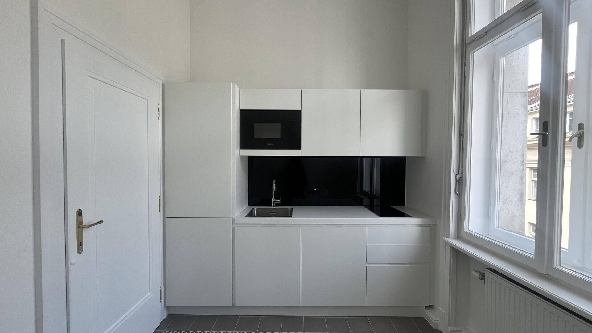 Moderne, kompakte Küchenzeile in Weiß mit integrierten Geräten und schwarzer Rückwand neben einem Fenster.
