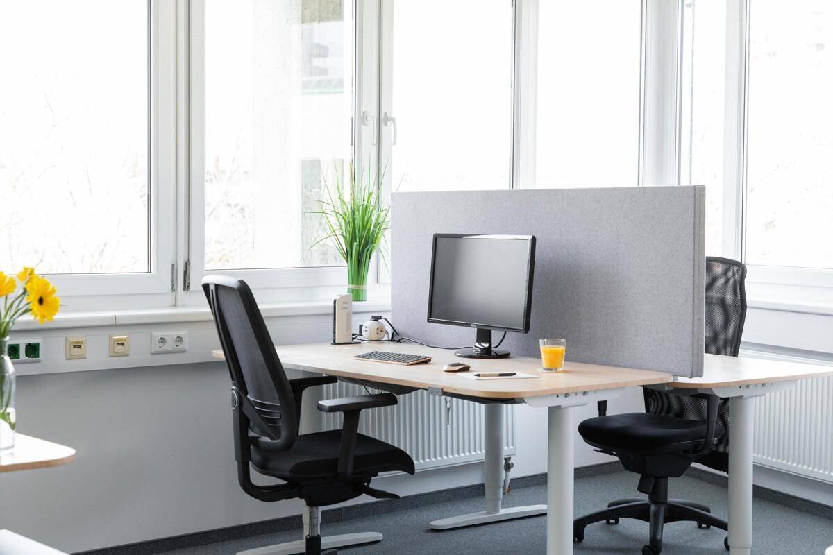Heller Arbeitsplatz mit ergonomischem Schreibtisch, Monitor und Bürostuhl vor großen Fenstern mit viel Tageslicht.