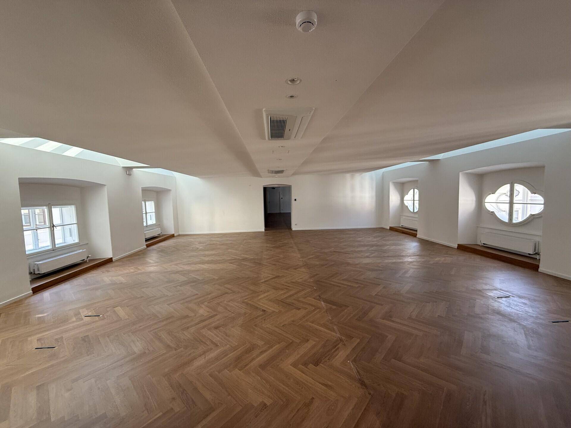 Weitläufiger Raum mit elegantem Fischgrätparkett, hohen Decken und mehreren Fenstern, die den Raum erhellen und Klimaanlage.