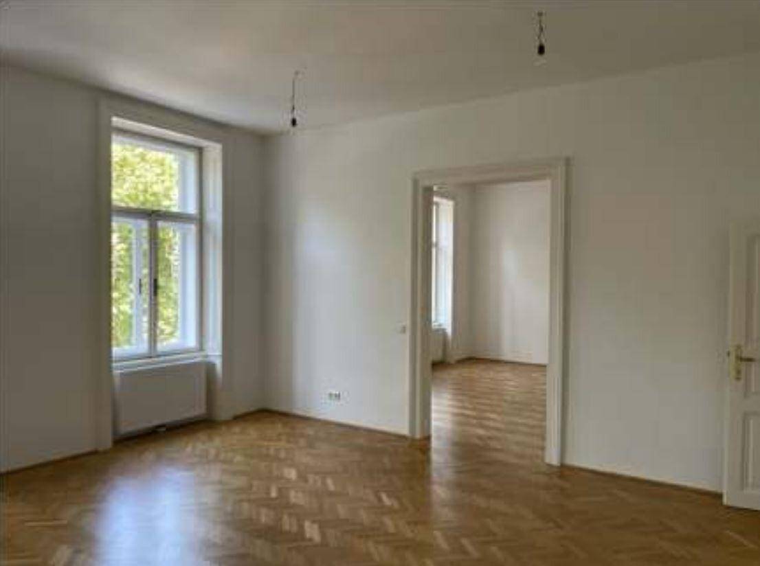 Geräumiger Raum mit Fischgrätparkett und hohen Fenstern, der zu einem weiteren Zimmer führt.