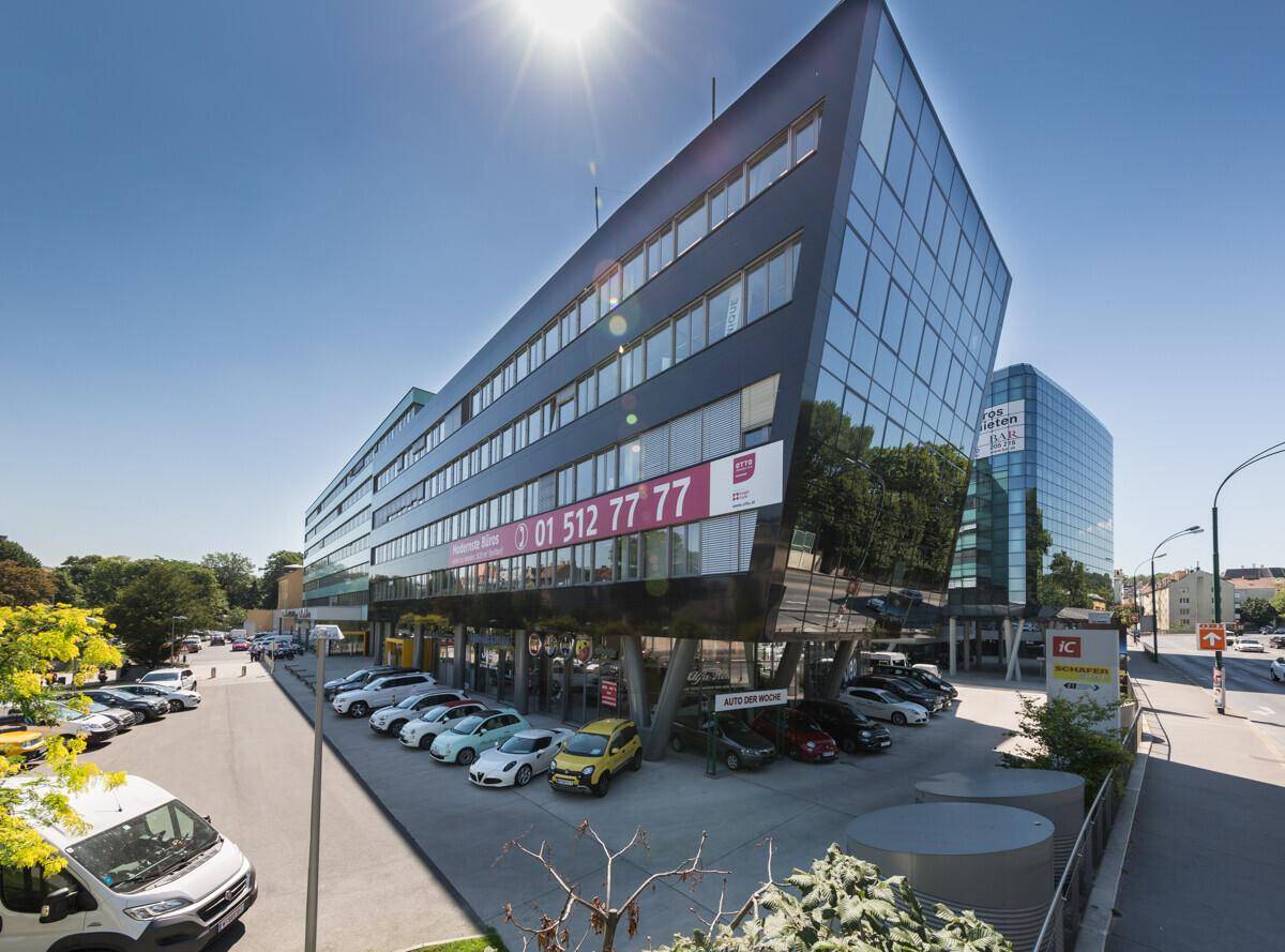 Weitwinkelaufnahme des modernen Bürogebäudes mit reflektierender Glasfassade und belebtem Parkbereich an einem sonnigen Tag.