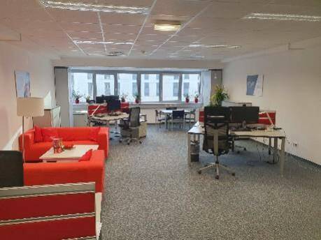 Geräumiger Büroraum mit mehreren Arbeitsplätzen, bequemen roten Sofas und großen Fenstern für viel Tageslicht.