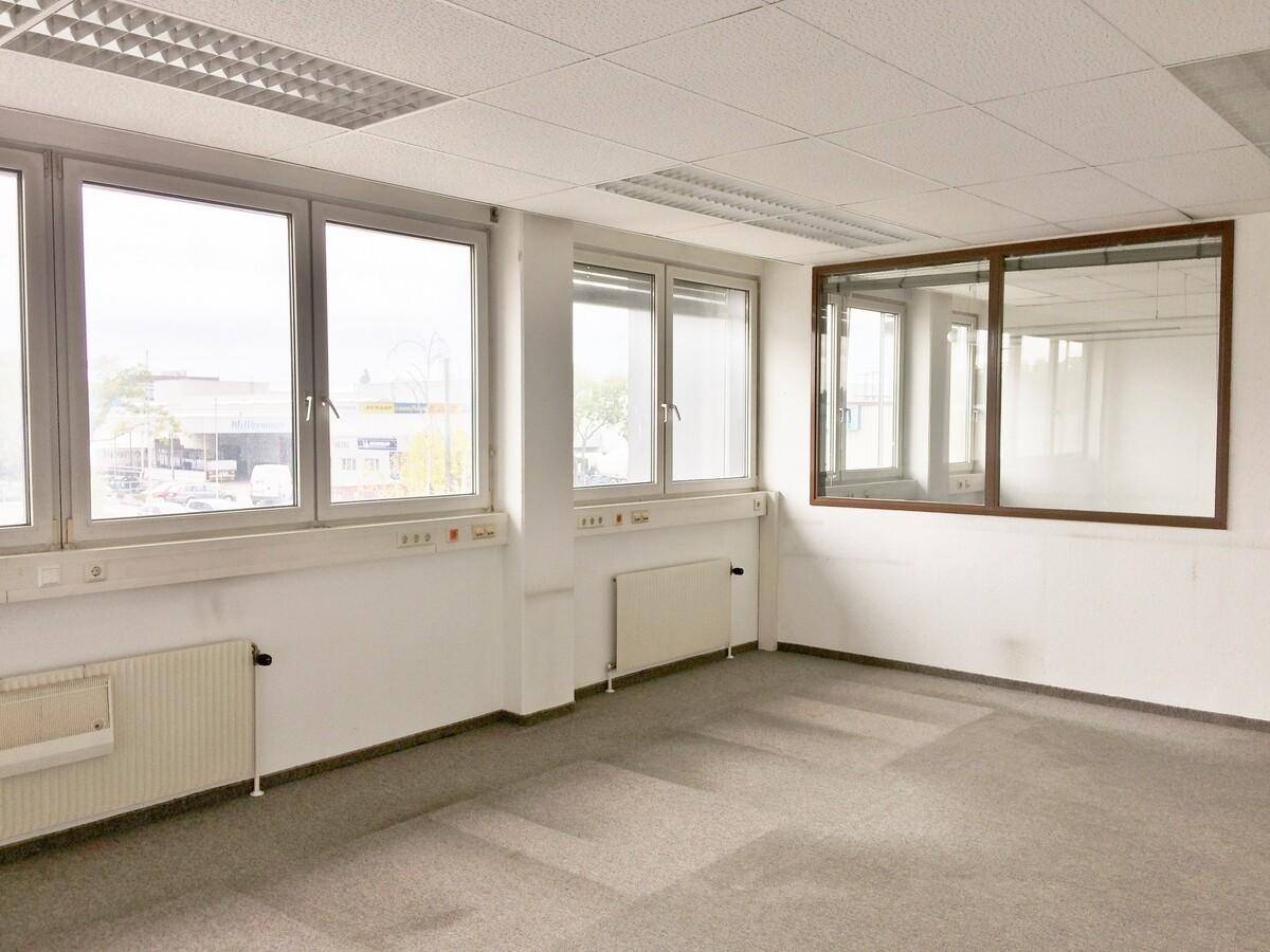 Helles Büro mit Teppichboden, großen Fenstern und einer internen Glaswand, die den Raum unterteilt.