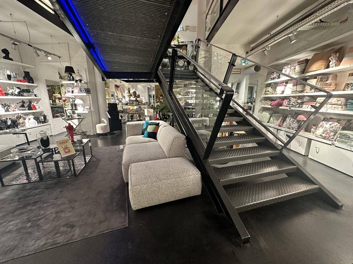 Verkaufsraum mit einer modernen Industrieltreppe aus Metall und Glas, umgeben von Möbeln.