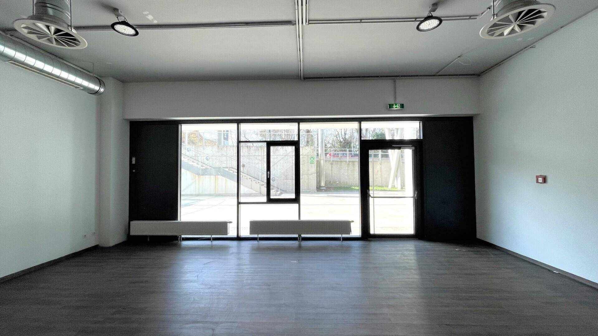 Heller, leerer Gewerberaum mit großen Fensterfronten, sichtbaren Lüftungsrohren an der Decke und dunklem Boden.