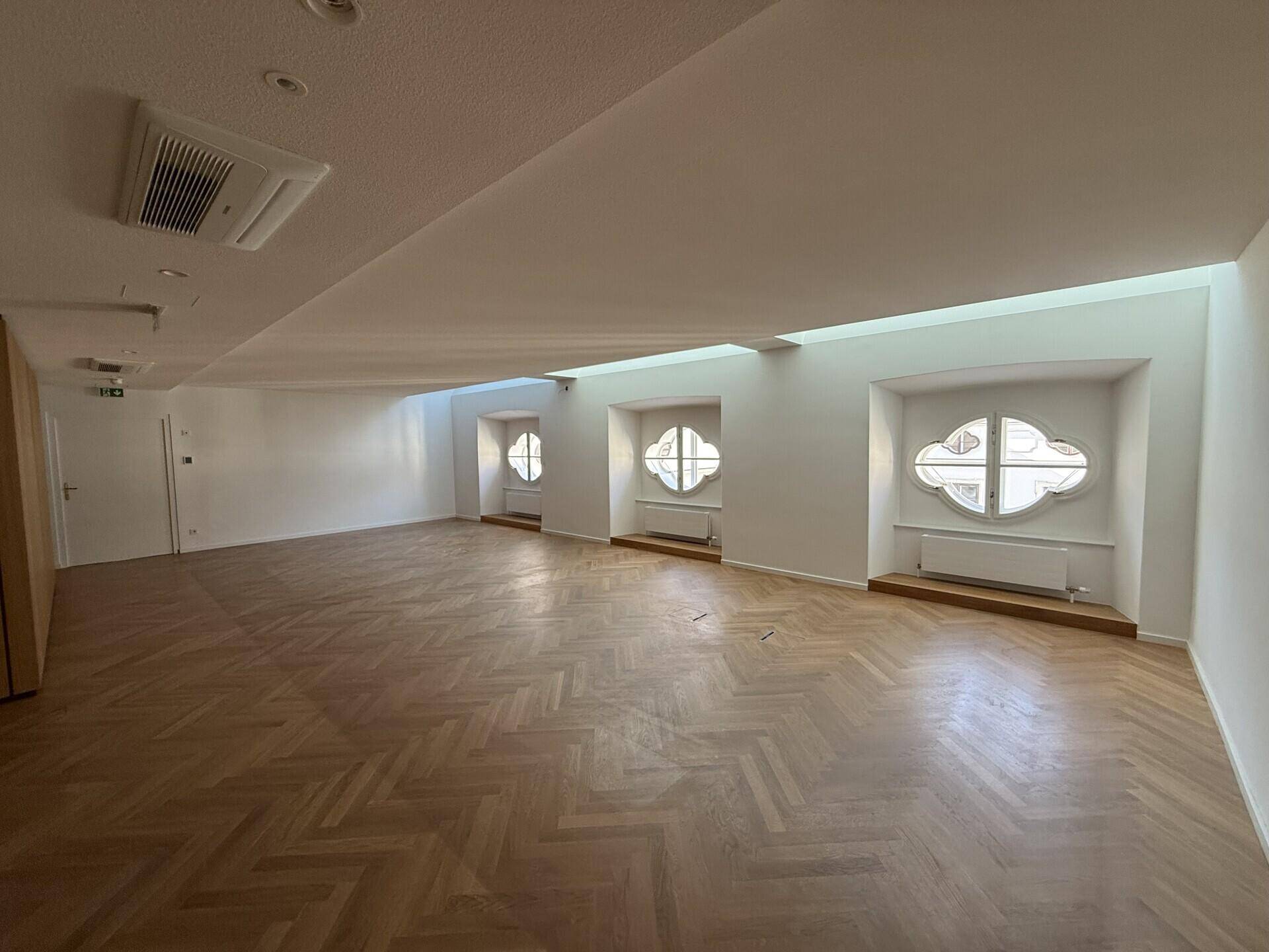 Heller, leerer Raum mit Fischgrätparkett, weißen Wänden und einzigartigen runden Fenstern, die Sonnenlicht auf den Boden werfen.