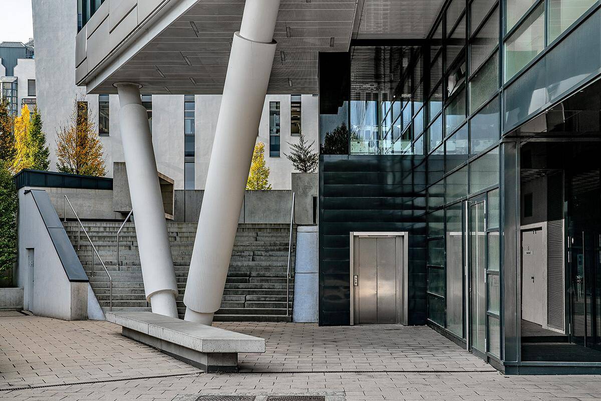 Eingangsbereich des Gebäudes mit markanten schrägen Säulen, einer Treppe und einem modernen Aufzug, der den Zugang erleichtert.