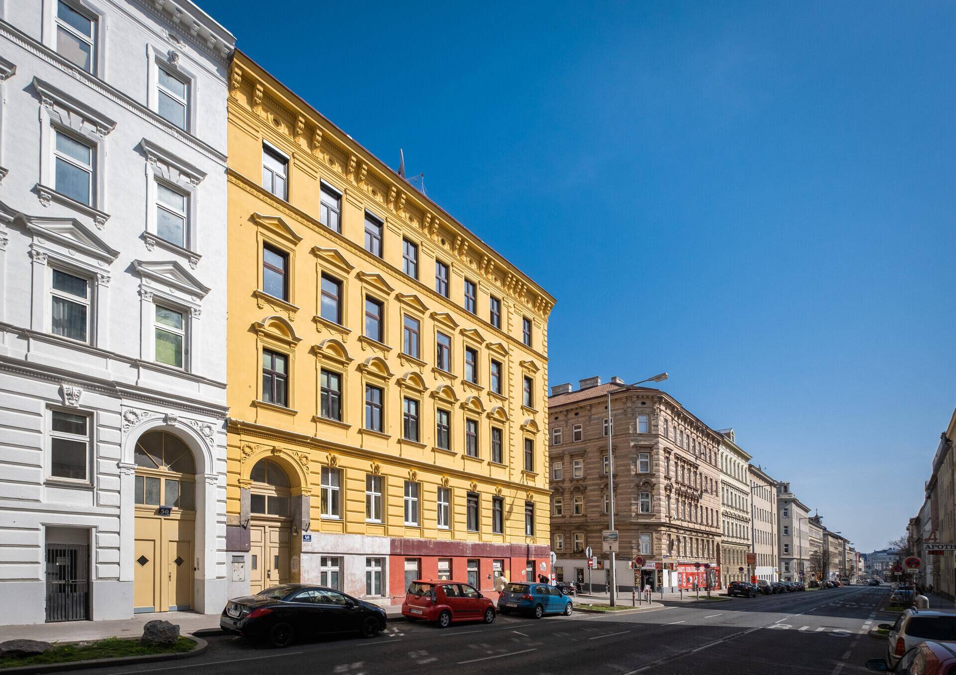 Seitenansicht eines historischen gelben Gebäudes an einer Straße mit parkenden Autos und klarem Himmel.