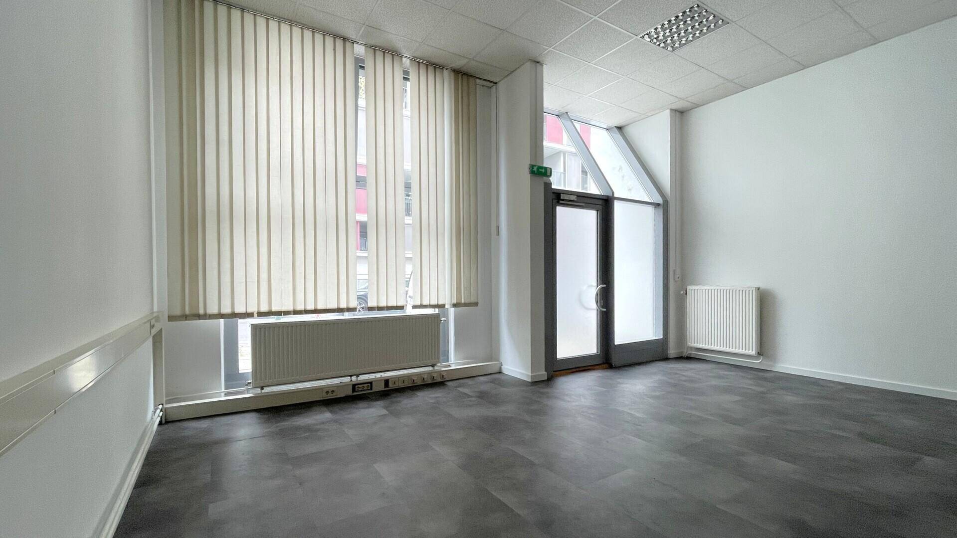 Heller Büroraum mit großen Fenstern, vertikalen Jalousien, Heizkörpern und dunklem Vinylboden.