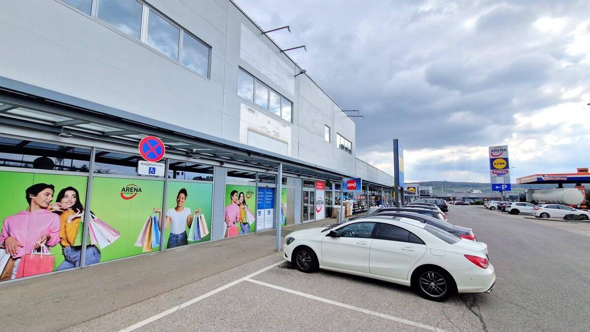 Außenansicht eines modernen Einkaufszentrums mit Parkplätzen und bewölktem Himmel.