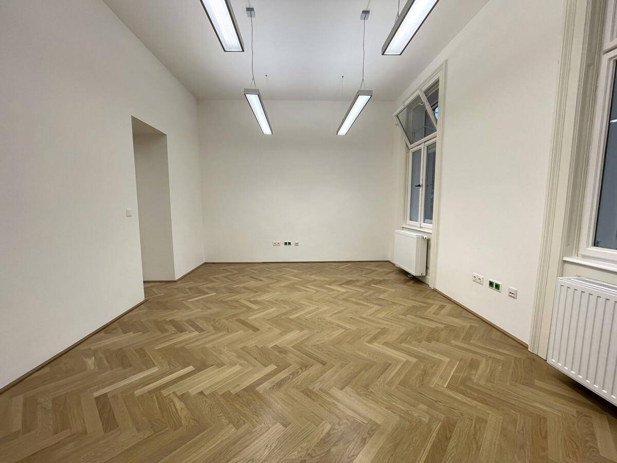 Heller Büroraum mit Fischgrätparkett und einem großen Fenster, das viel Tageslicht hereinlässt.