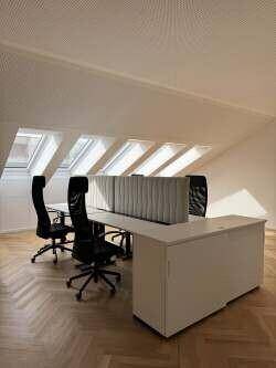 Helles Büro mit langen Schreibtischen, Bürostühlen und Fischgrätparkett unter schrägen Dachfenstern.
