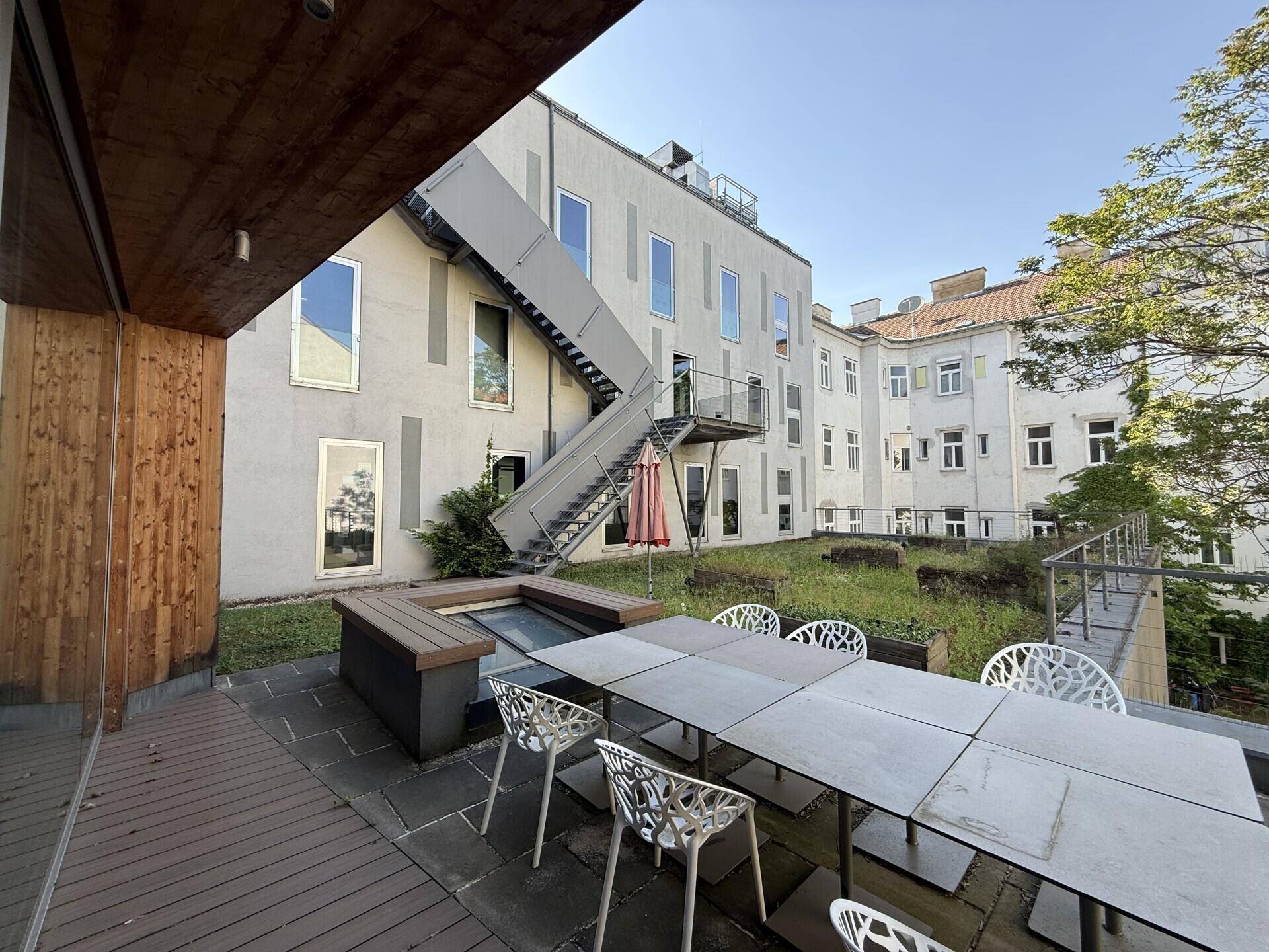 Geräumige Terrasse mit Holzboden und Sitzgelegenheiten, Blick auf das Gebäude mit Außentreppe.
