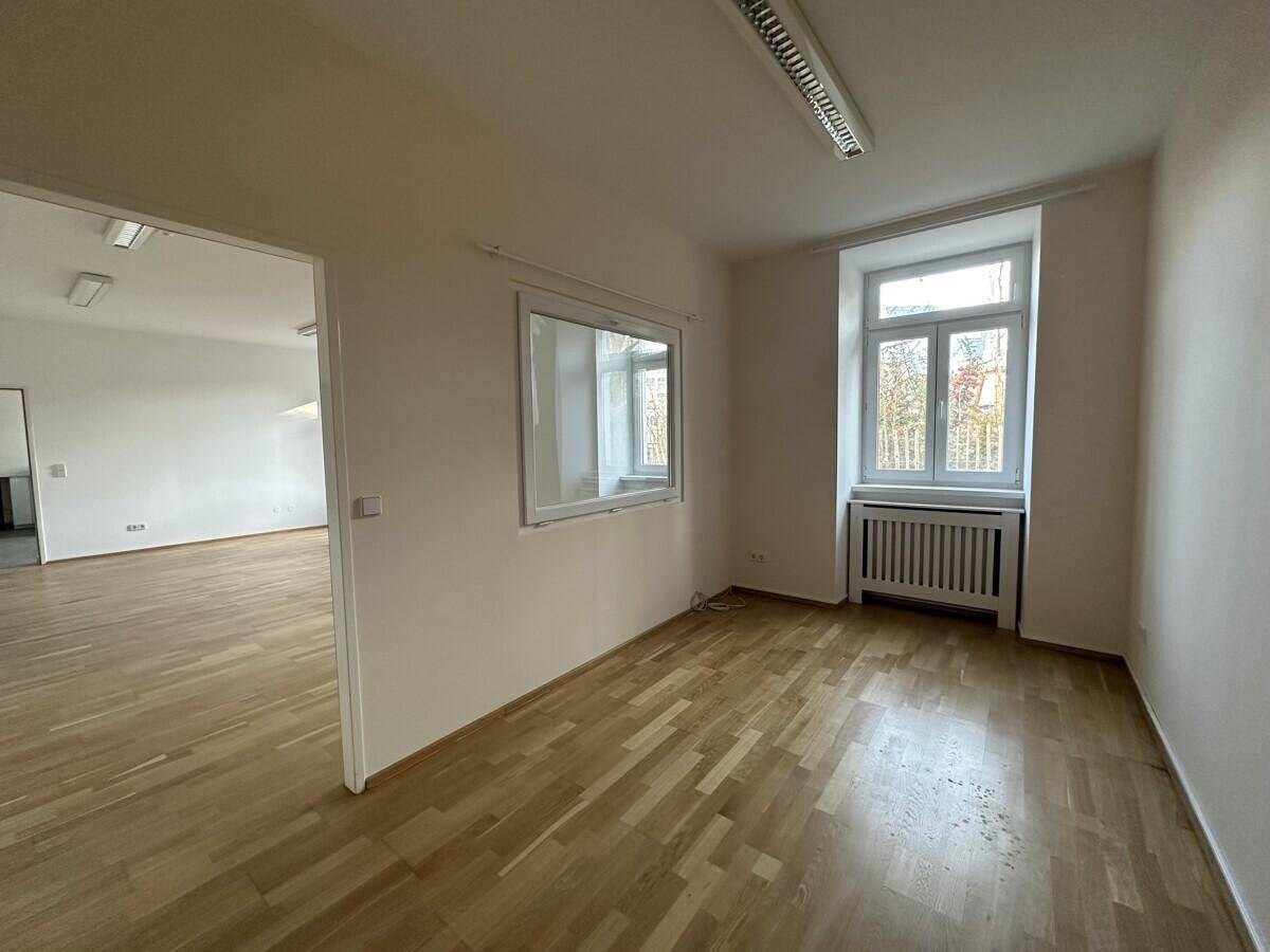Büroraum mit Parkettboden, einem Fenster und einer internen Fensteröffnung zu einem anderen Bereich.