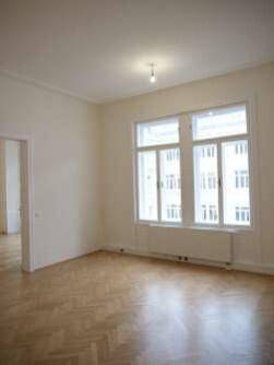 Helles Zimmer mit großen Fenstern, Parkettboden und Blick auf die gegenüberliegenden Gebäude.