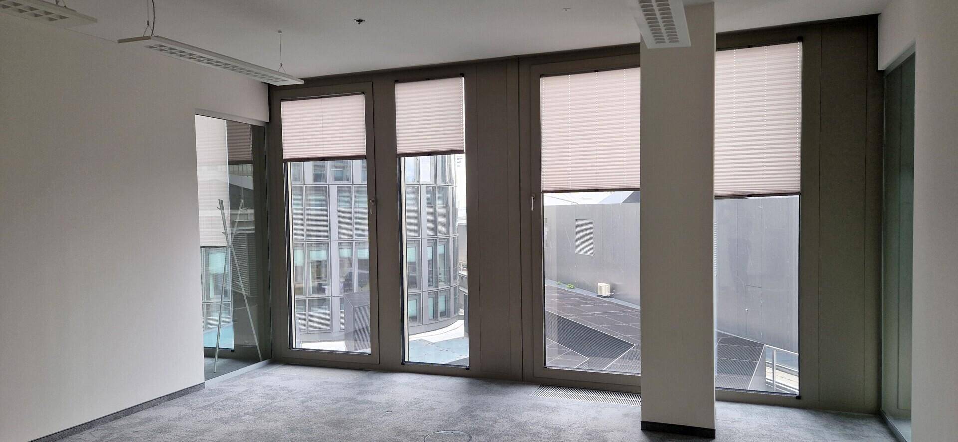 Leerer Büroraum mit großen Fenstern, Jalousien und Blick auf umliegende Gebäude.