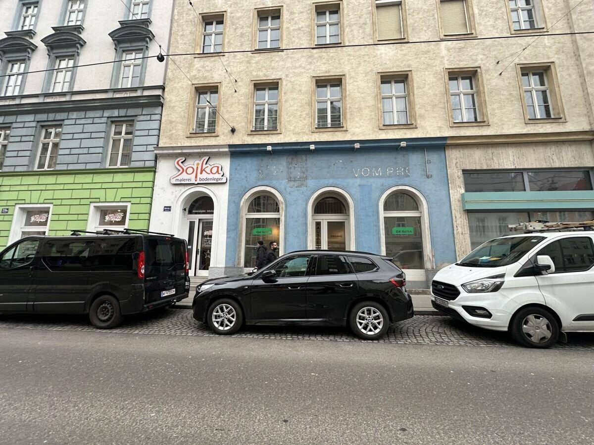 Straßenansicht eines Gebäudes mit Geschäften im Erdgeschoss und parkenden Autos davor unter bewölktem Himmel.