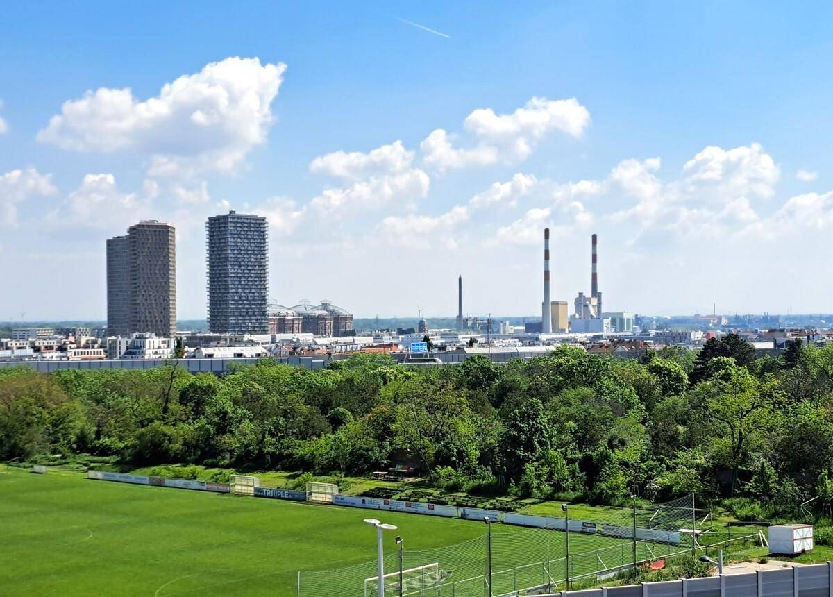 Weitläufiger Blick über eine Stadt mit Sportplatz, Bäumen und Industrieanlagen im Hintergrund unter blauem Himmel.