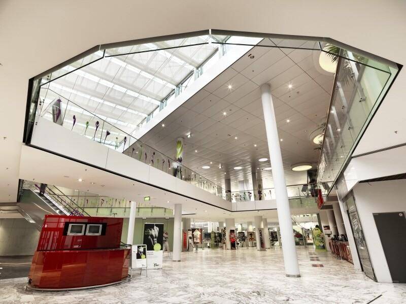 Geräumiges, modernes Atrium mit Glasdach, Rolltreppen und einem auffälligen roten Empfangstresen auf Marmorboden.