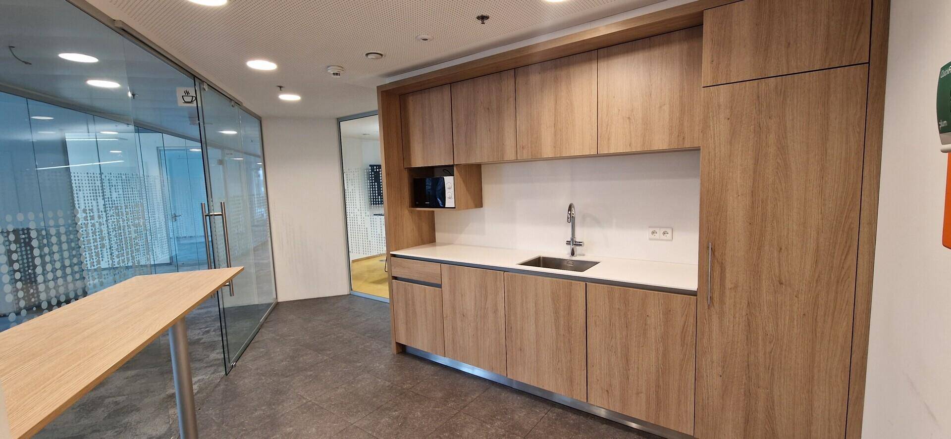 Funktionale Teeküche mit hellen Holzschränken und integrierter Mikrowelle, angrenzend an einen Glasbürobereich.