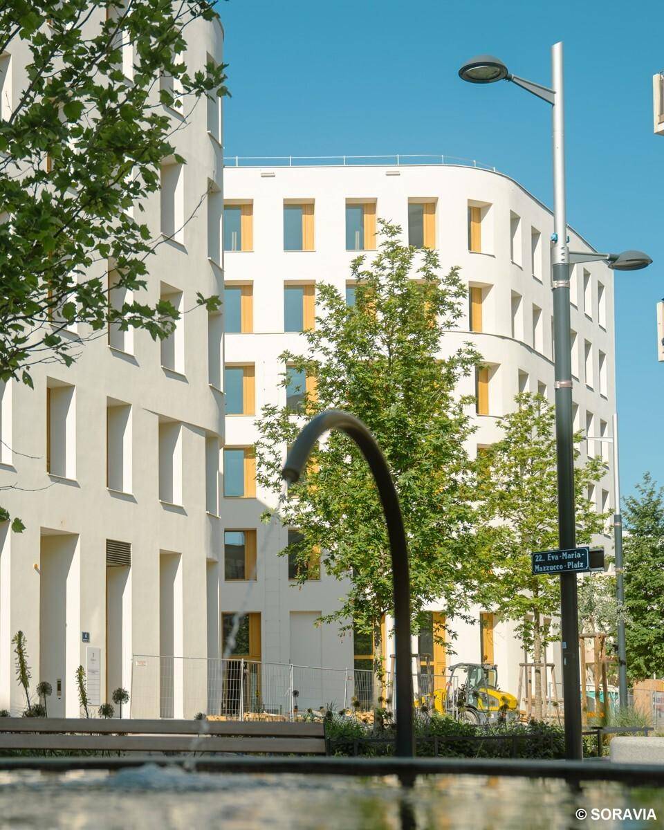 Moderne weiße Gebäude umgeben von Bäumen und Straßenlaternen, mit einer Baustelle im Hintergrund.