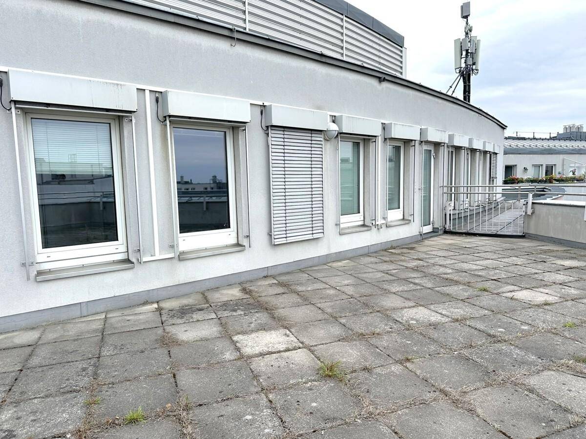 Teil einer Dachterrasse mit Betonplatten und Fenstern mit Rollläden an der Gebäudefassade.