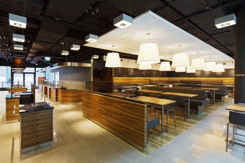 Großzügige, moderne Cafeteria mit Holzakzenten, vielfältigen Sitzgelegenheiten und einer Essensausgabe.