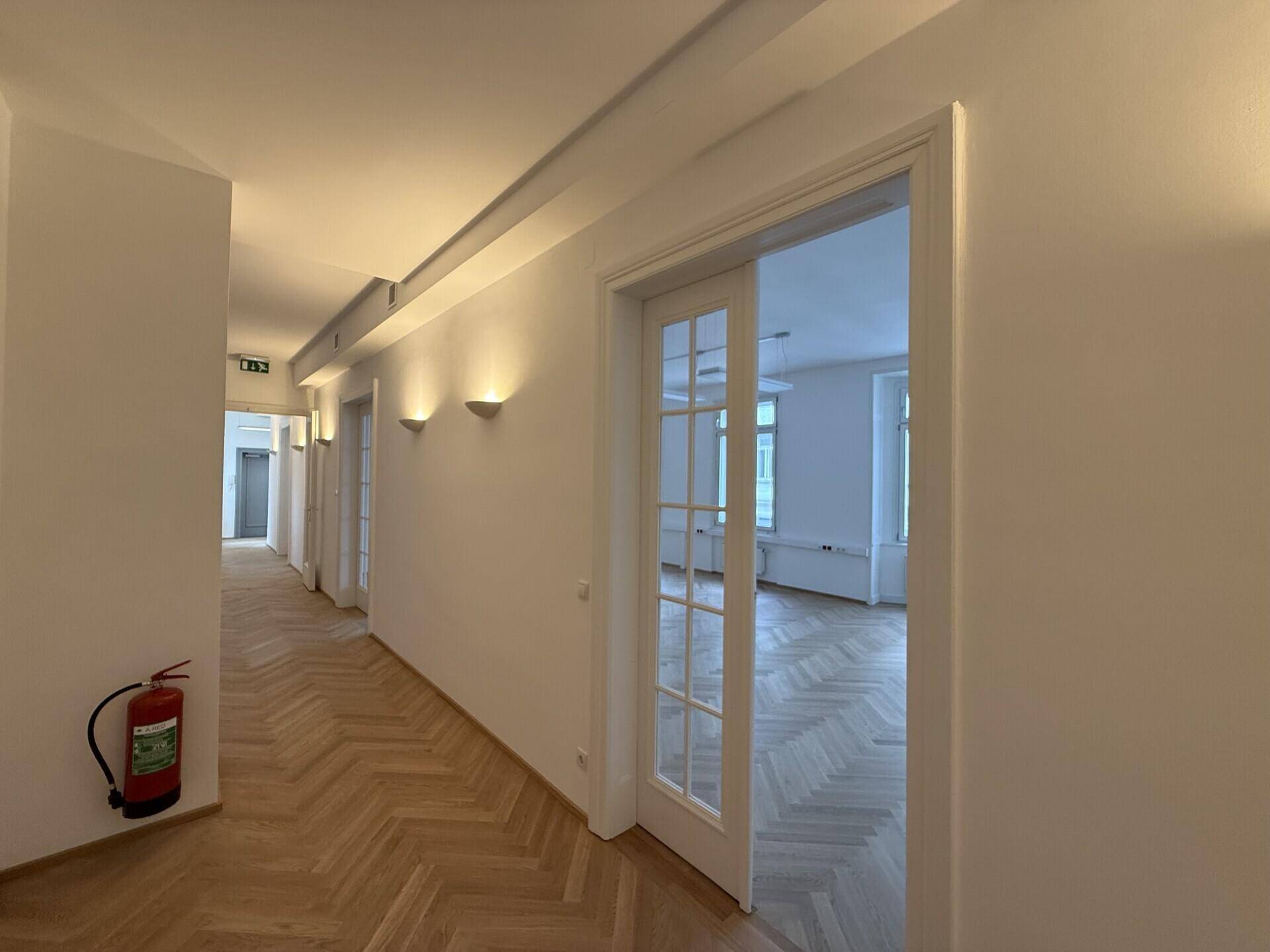 Langer Flur mit Fischgrätparkett, Wandleuchten und einem Feuerlöscher an der Wand, gut beleuchtet.