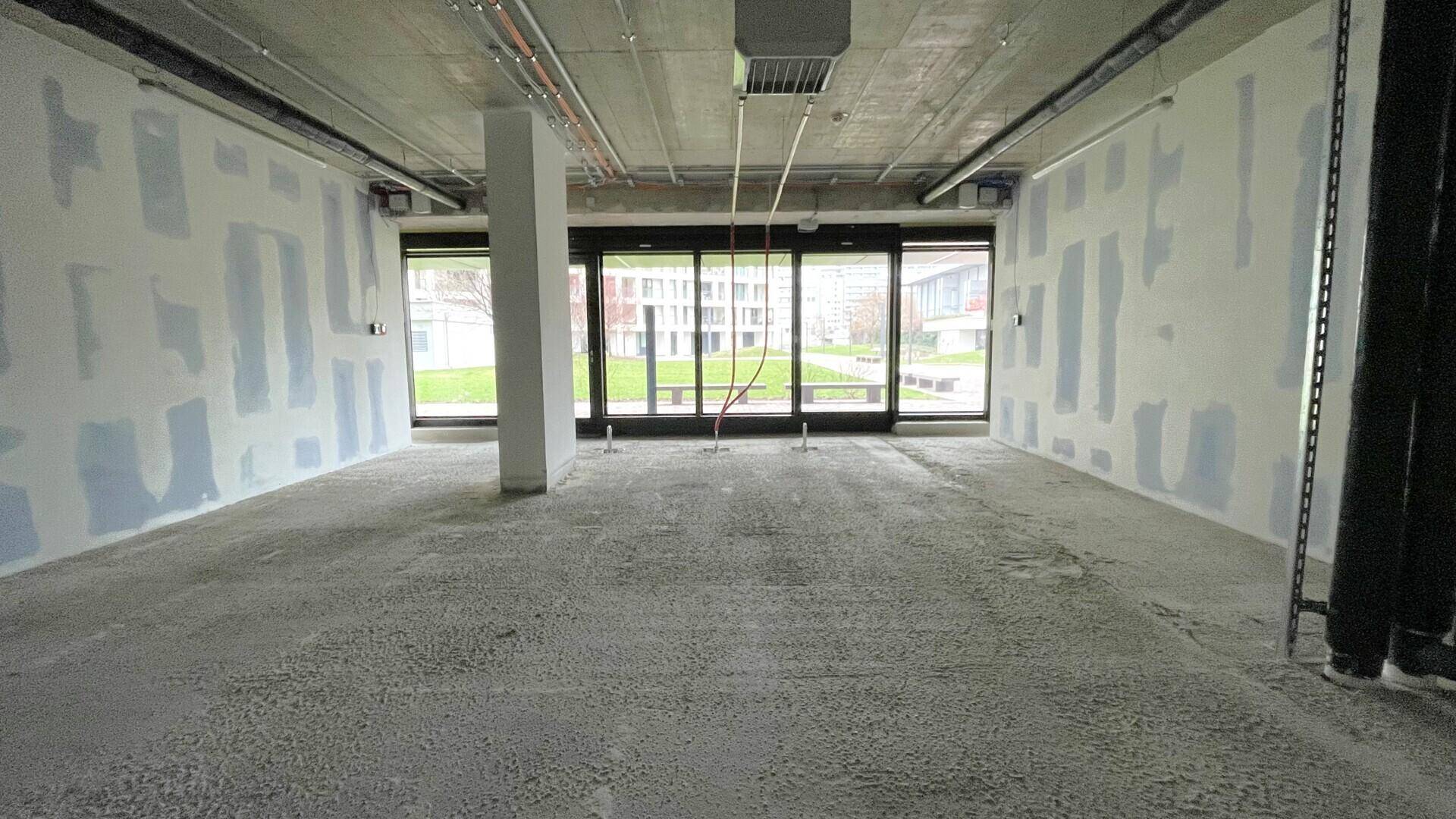 Heller Gewerberaum im Rohbauzustand mit großen Fenstern, die viel Tageslicht hereinlassen und einen Blick ins Grüne bieten.