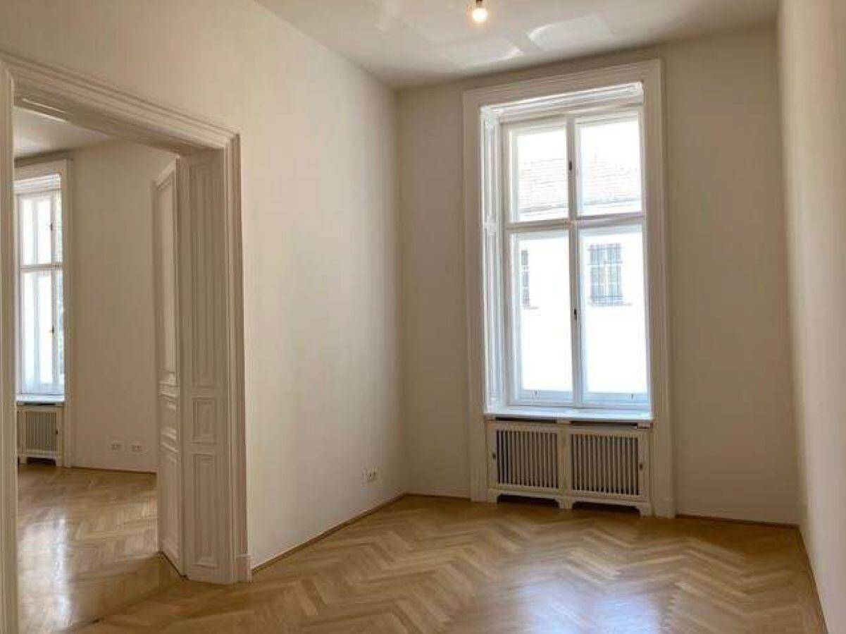 Heller Wohnraum mit Fischgrätparkett, weißen Wänden und einem Durchgang zu einem weiteren Zimmer.