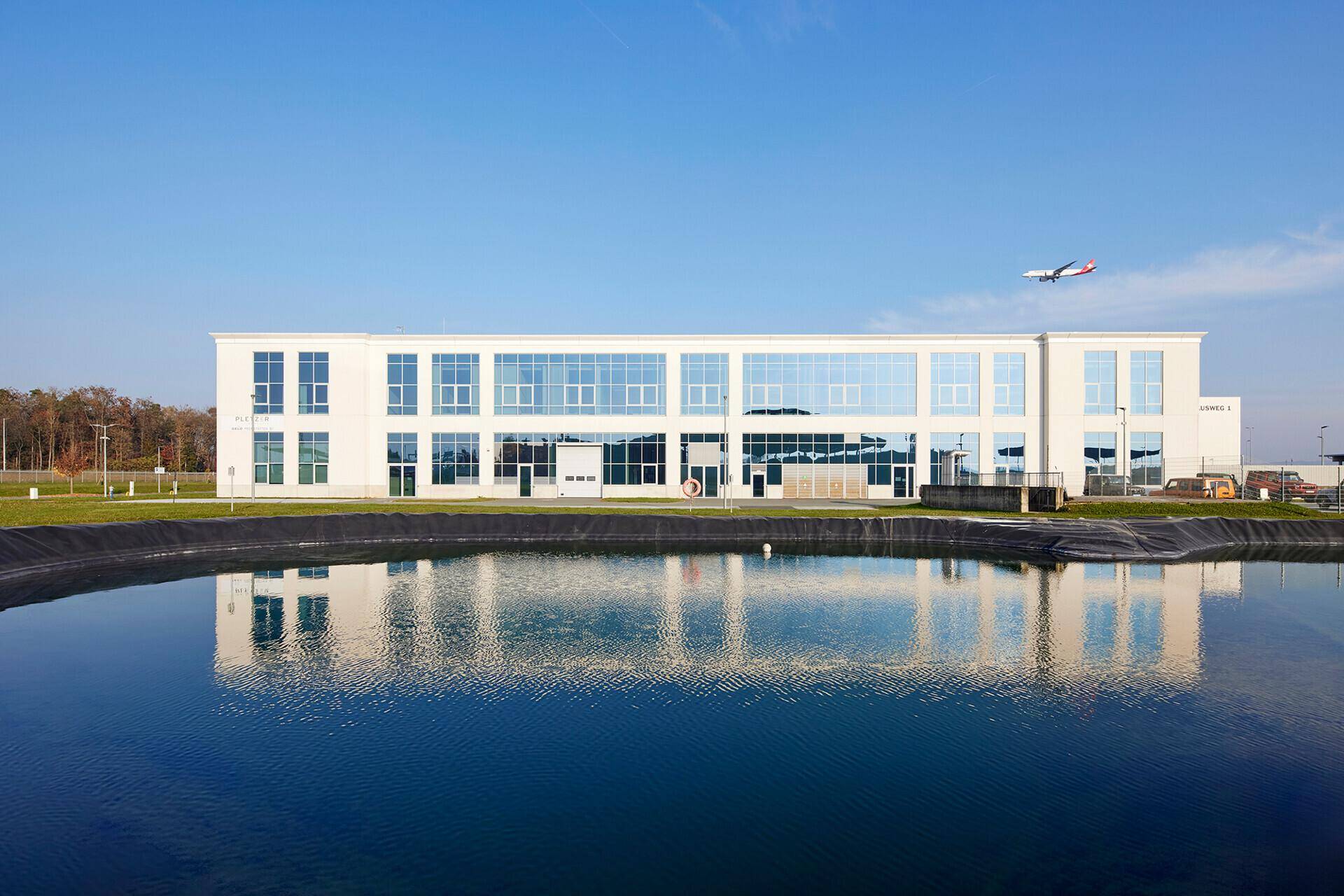 Modernes Geschäftsgebäude spiegelt sich in einem Wasserbecken wider, mit einem Flugzeug am klaren Himmel.