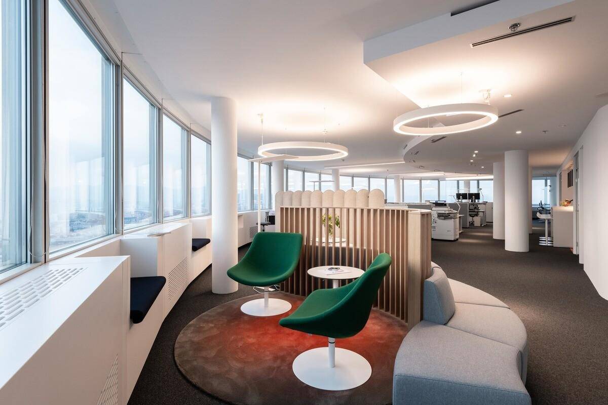 Moderner Bürobereich mit grünen Sesseln, runden Sofas und einer Holzabtrennung, umgeben von großen Fenstern.