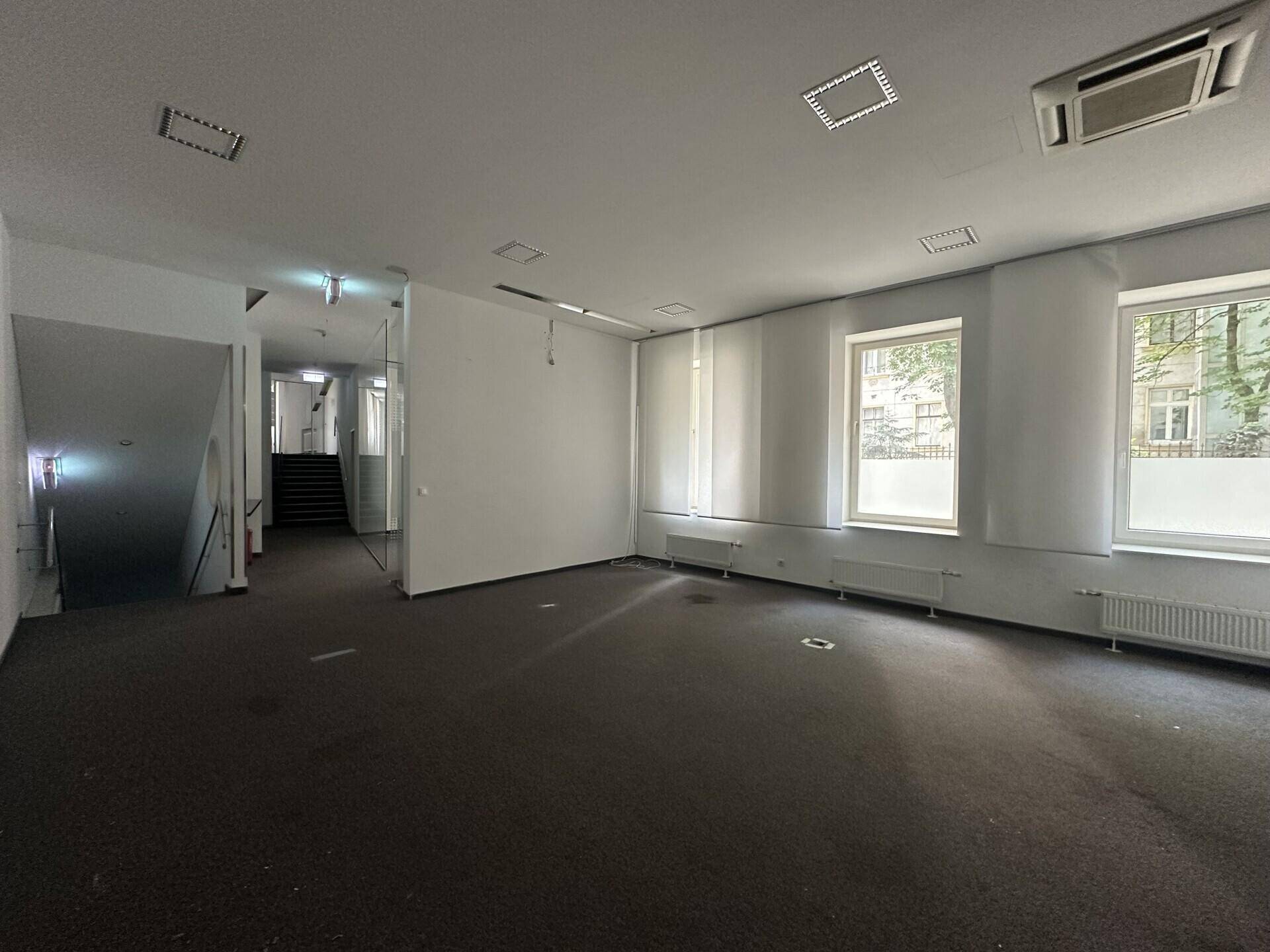 Geräumiger Büroraum mit Teppichboden, Fenstern und einem sichtbaren Treppenaufgang im Hintergrund.