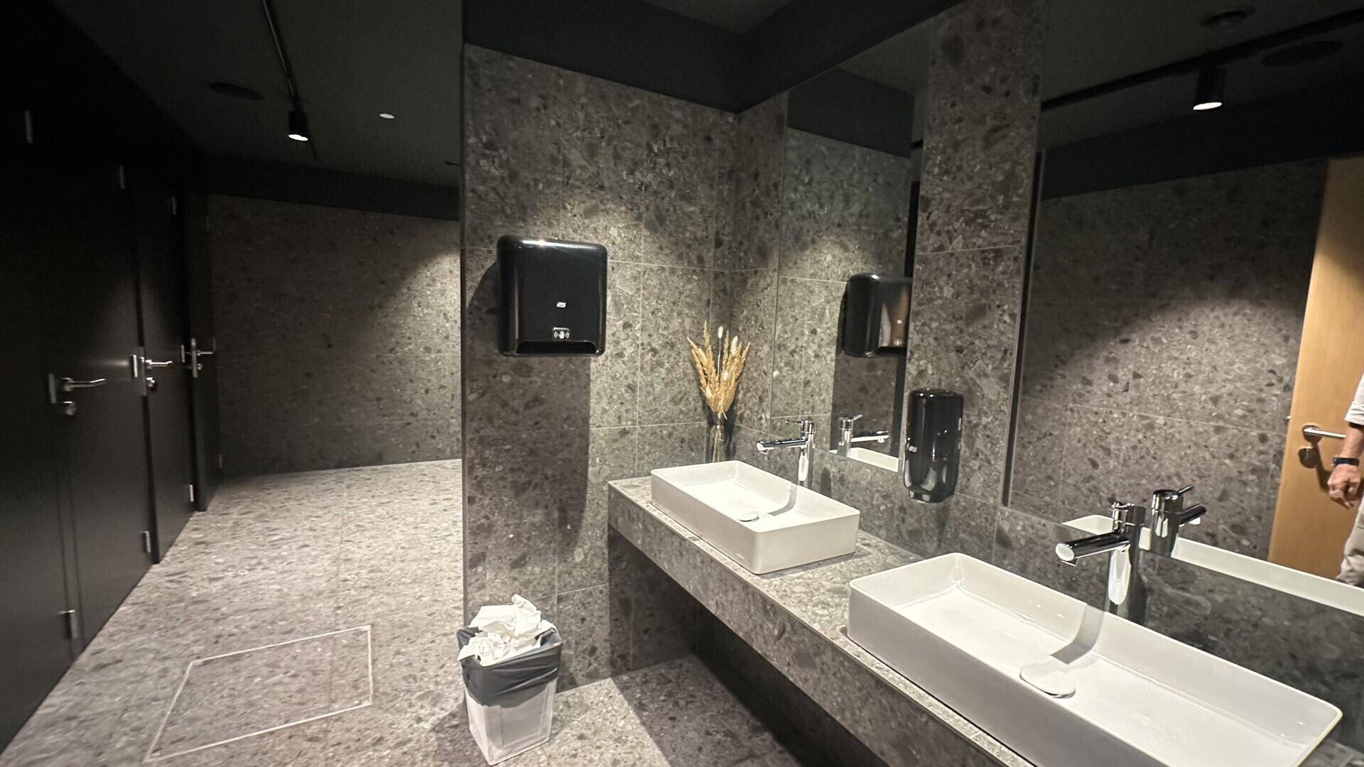 Moderner Waschraum mit zwei Waschbecken, großen Spiegeln und grauen Steinfliesen, in minimalistischem Design.