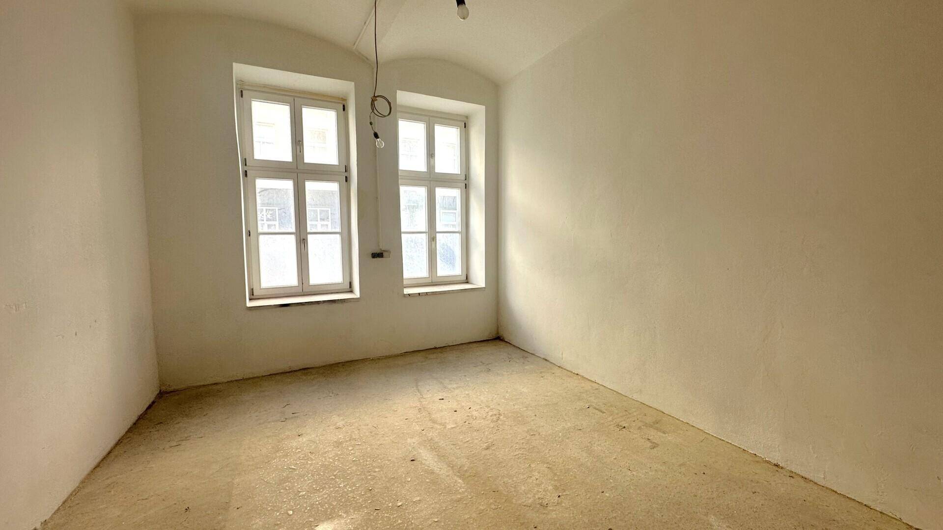 Ein leerer Raum mit zwei hohen Fenstern, die viel Tageslicht hereinlassen, und einem einfachen Betonboden.
