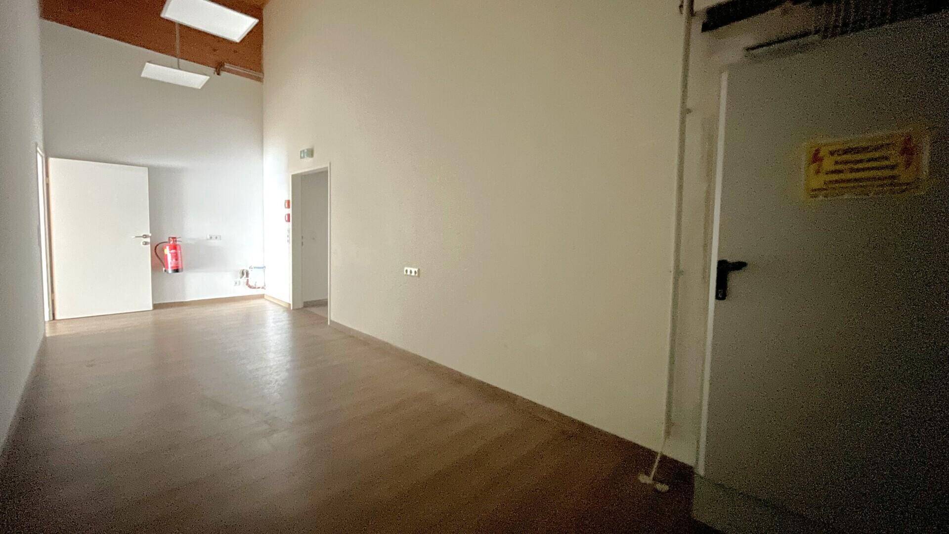 Langer Flur mit Holzbalkendecke, hellem Boden und mehreren Türen, darunter ein Notausgang und ein Feuerlöscher.