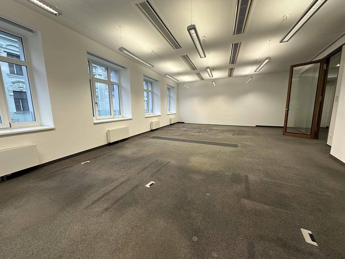 Geräumiger Büroraum mit Teppichboden, großen Fenstern und einer Glastür, bietet viel Licht und Offenheit.
