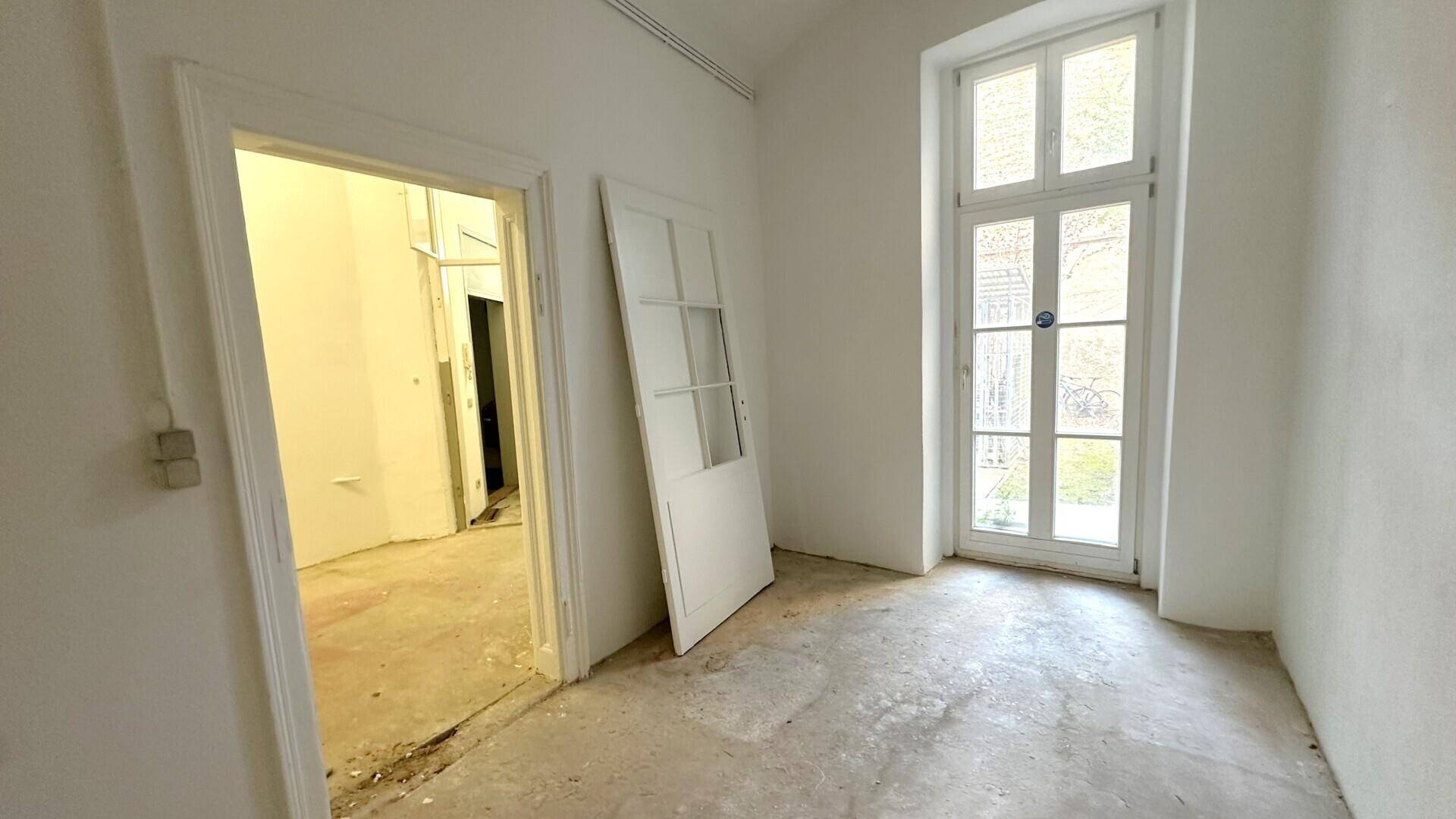 Ein heller Raum mit einer hohen Fenstertür, die nach draußen führt, und einer offenen Tür zu einem weiteren Raum.