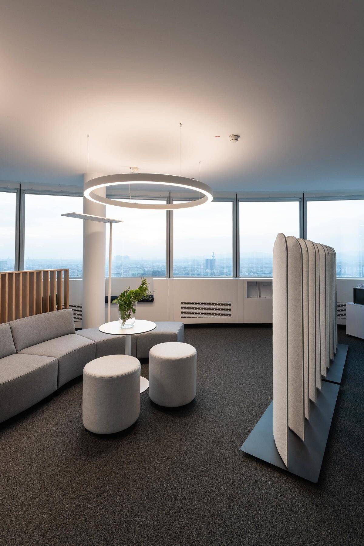 Moderner Bürobereich mit runden Sofas, Hockern und einer Trennwand, umgeben von großen Fenstern mit Stadtblick.