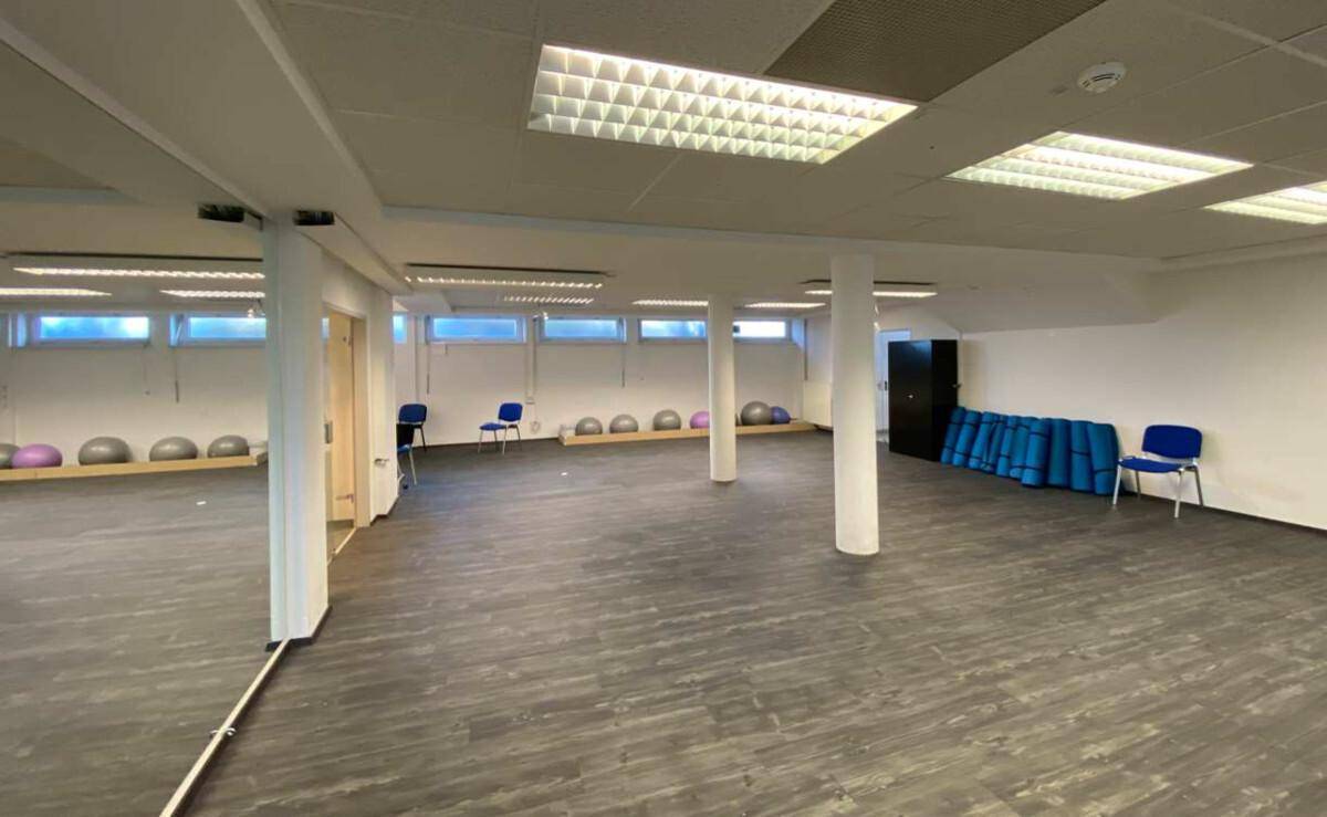 Großer Mehrzweckraum mit dunklem Vinylboden, tragenden Säulen und Sportgeräten an den Wänden.