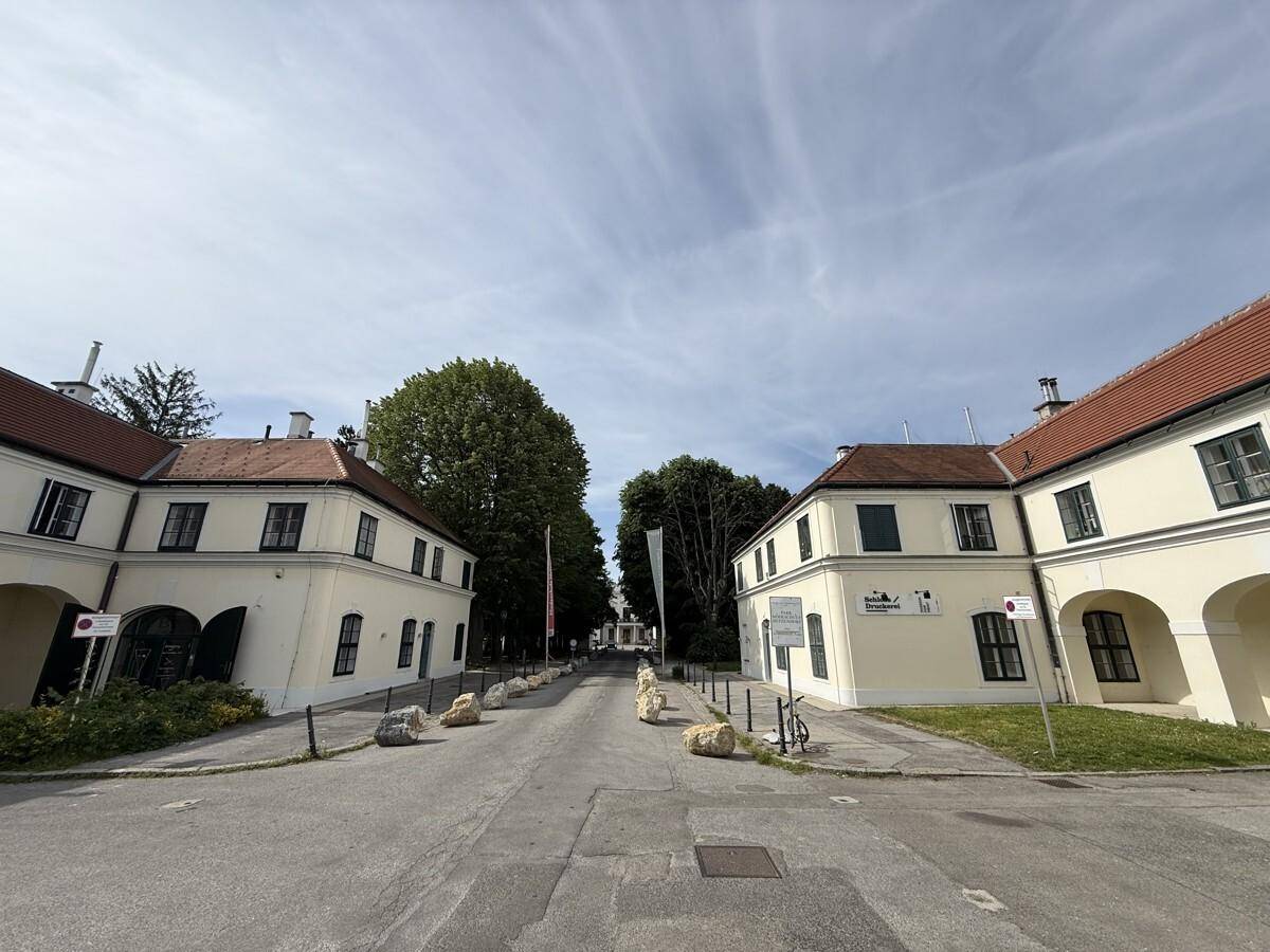 Blick auf zwei historische Gebäude mit hellen Fassaden und roten Dächern, die eine Allee säumen.