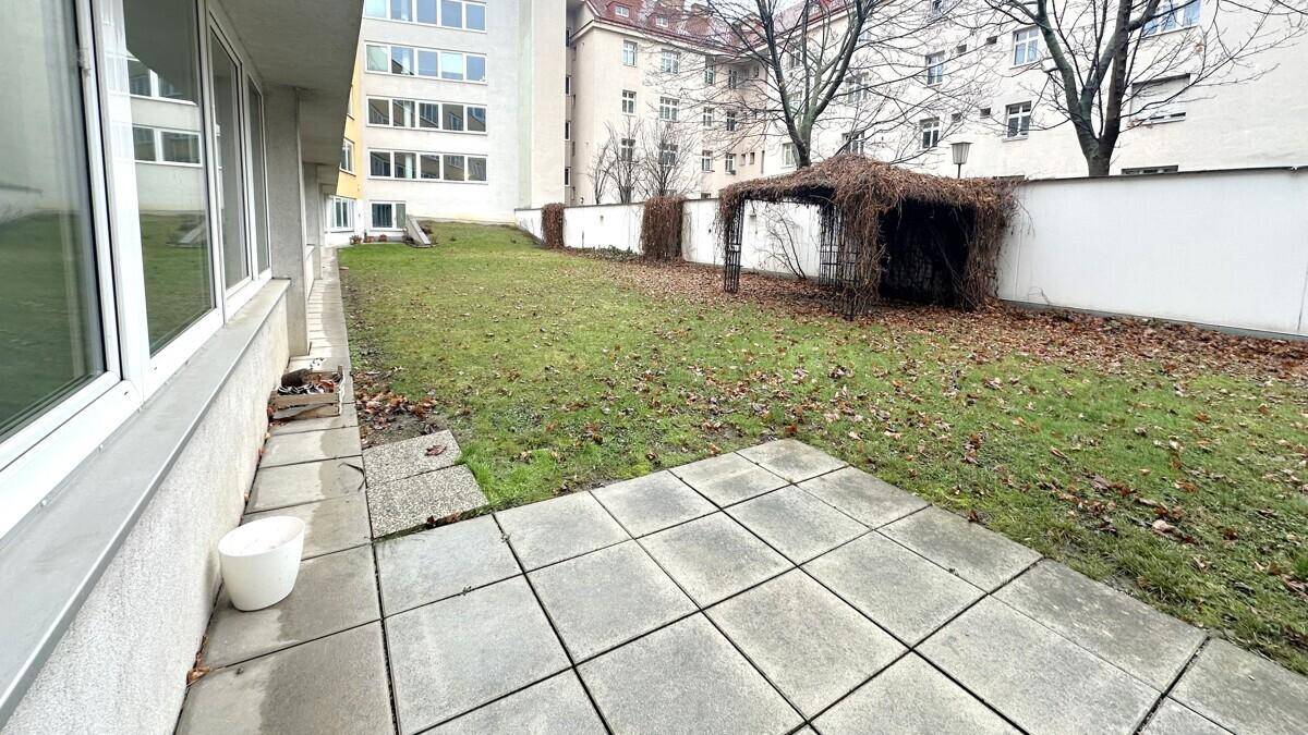 Kleiner Gartenbereich mit gepflasterter Terrasse, Rasenfläche und einem Rankgerüst im Hintergrund.
