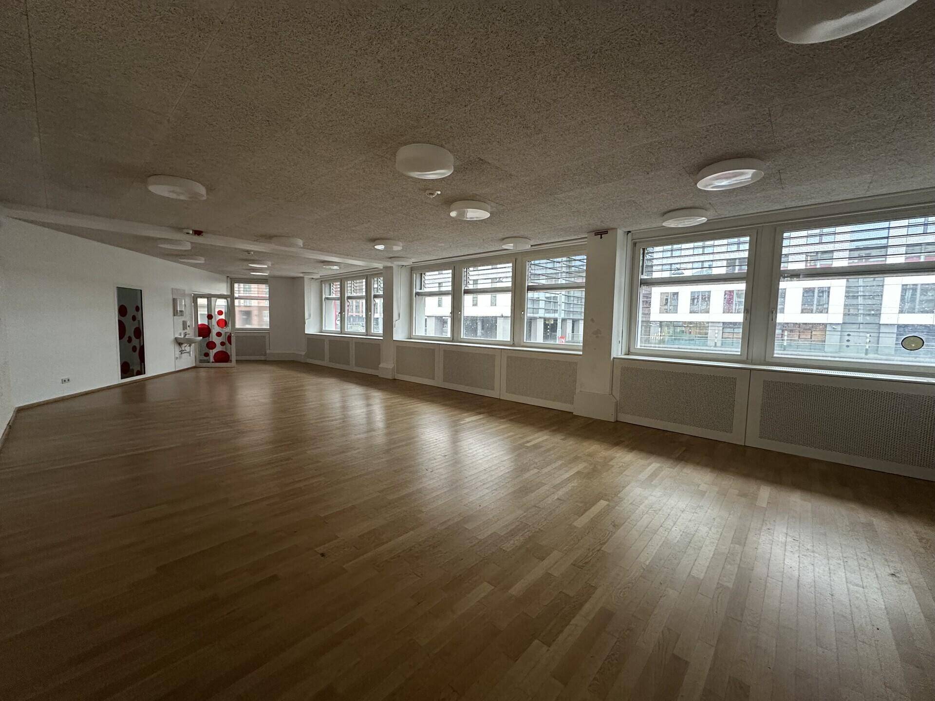 Heller und weitläufiger Raum mit Holzboden und einer Reihe großer Fenster, die viel Licht hereinlassen.