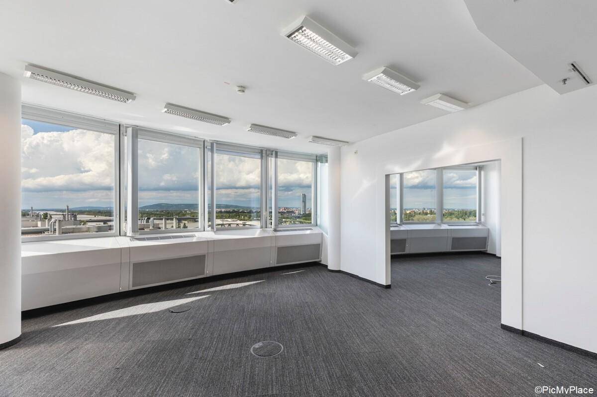 Geräumiger, leerer Büroraum mit Teppichboden, großen Fenstern und Blick auf die Stadt und die umliegende Landschaft.